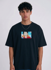 A full-body studio photograph of a male model wearing an Anetoonz black oversized crewneck t-shirt featuring a framed graphic print of a smiling Monkey D. Luffy with a straw hat against a colorful background.