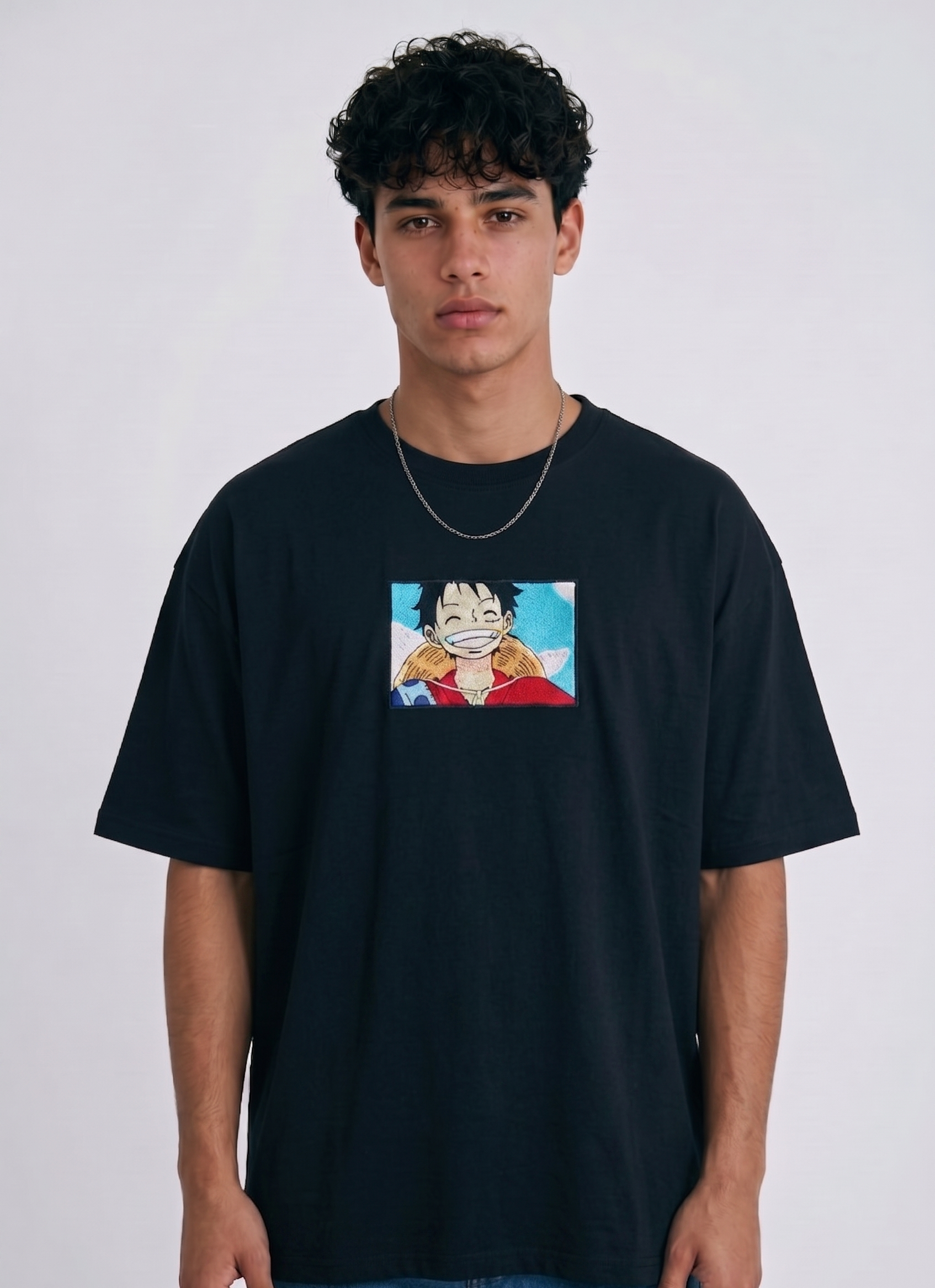 A full-body studio photograph of a male model wearing an Anetoonz black oversized crewneck t-shirt featuring a framed graphic print of a smiling Monkey D. Luffy with a straw hat against a colorful background.