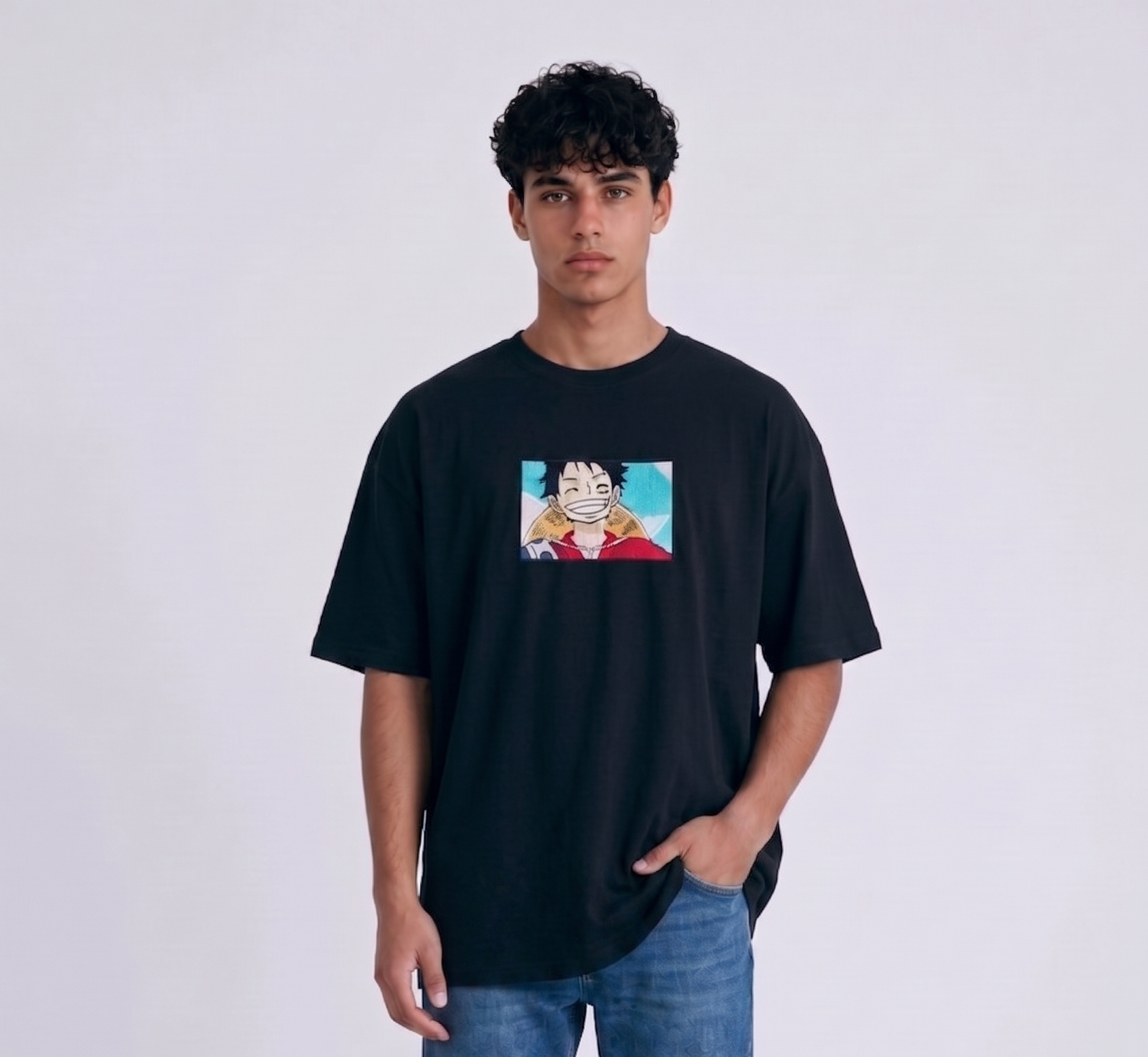 A full-body studio photograph of a male model wearing an Anetoonz black oversized crewneck t-shirt featuring a framed graphic print of a smiling Monkey D. Luffy with a straw hat against a colorful background.