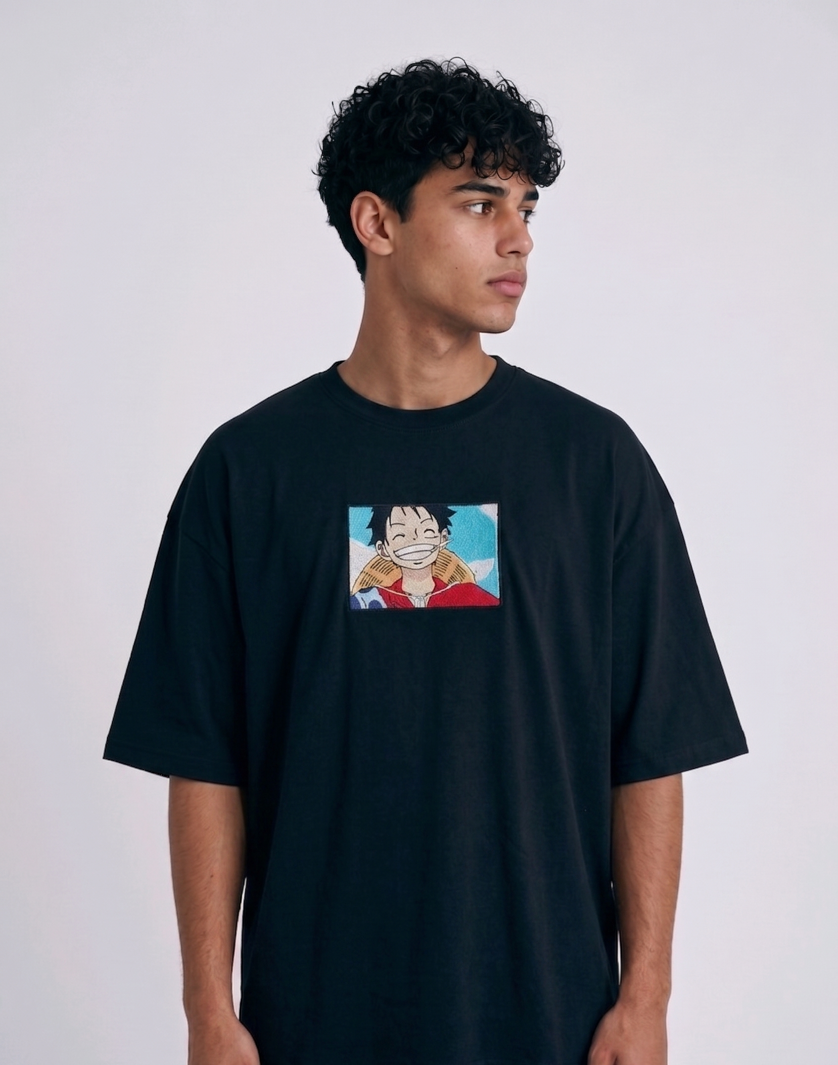 A full-body studio photograph of a male model wearing an Anetoonz black oversized crewneck t-shirt featuring a framed graphic print of a smiling Monkey D. Luffy with a straw hat against a colorful background.