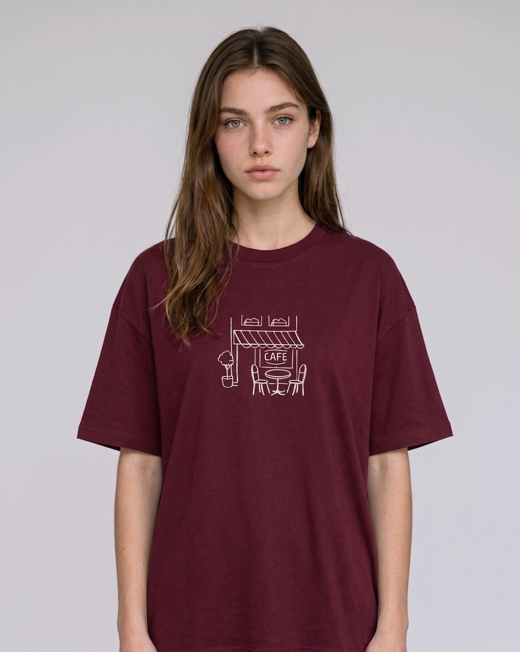 Wearing a cream-colored, crew-neck t-shirt. The center of the chest features a minimalist black line-art embroidery depicting a classic cafe storefront.

