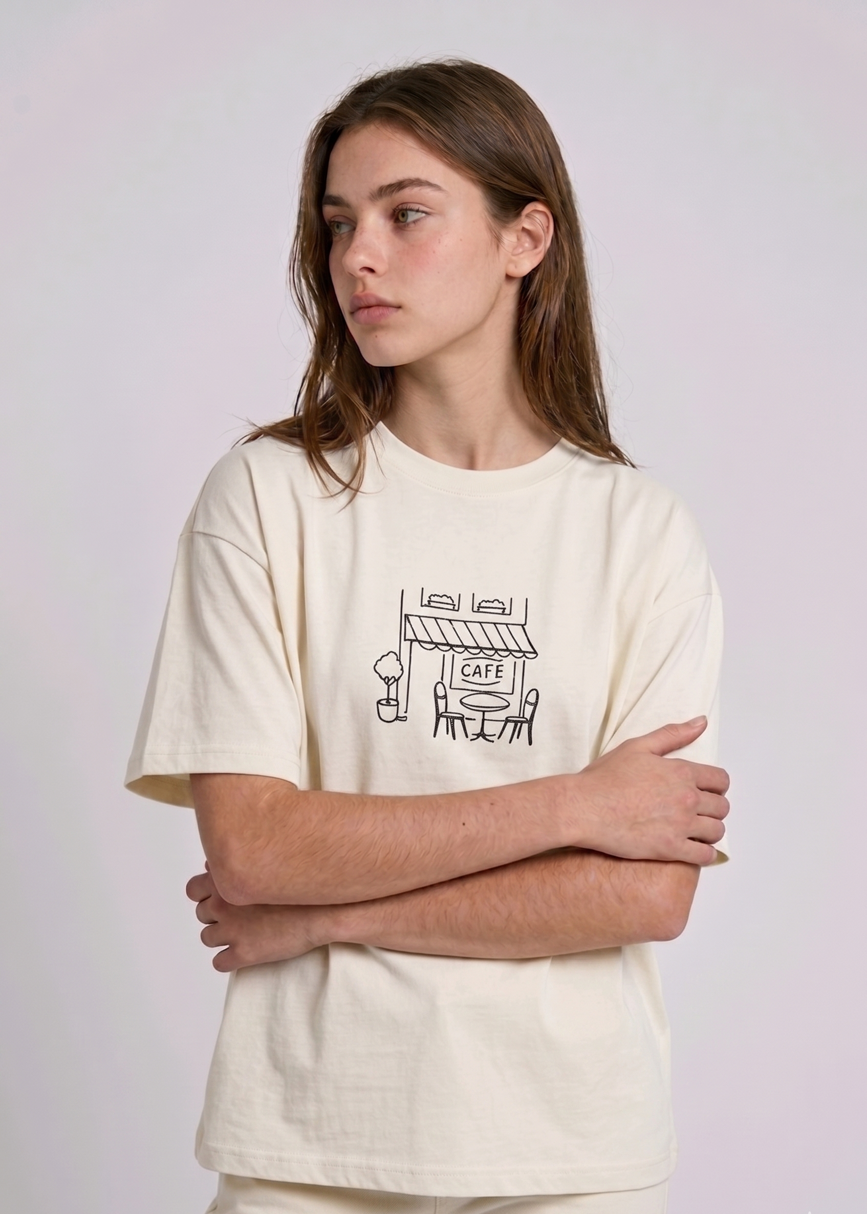 Wearing a cream-colored, crew-neck t-shirt. The center of the chest features a minimalist black line-art embroidery depicting a classic cafe storefront.

