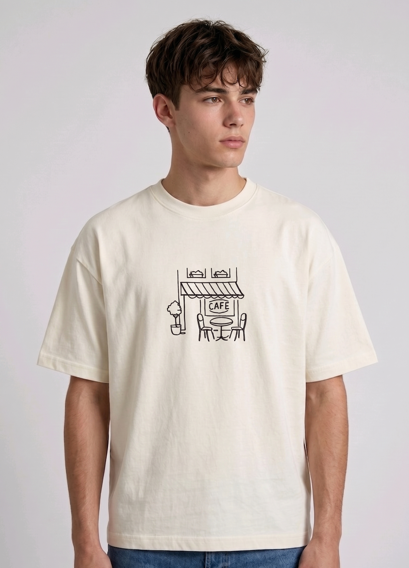 Wearing a cream-colored, crew-neck t-shirt. The center of the chest features a minimalist black line-art embroidery depicting a classic cafe storefront.

