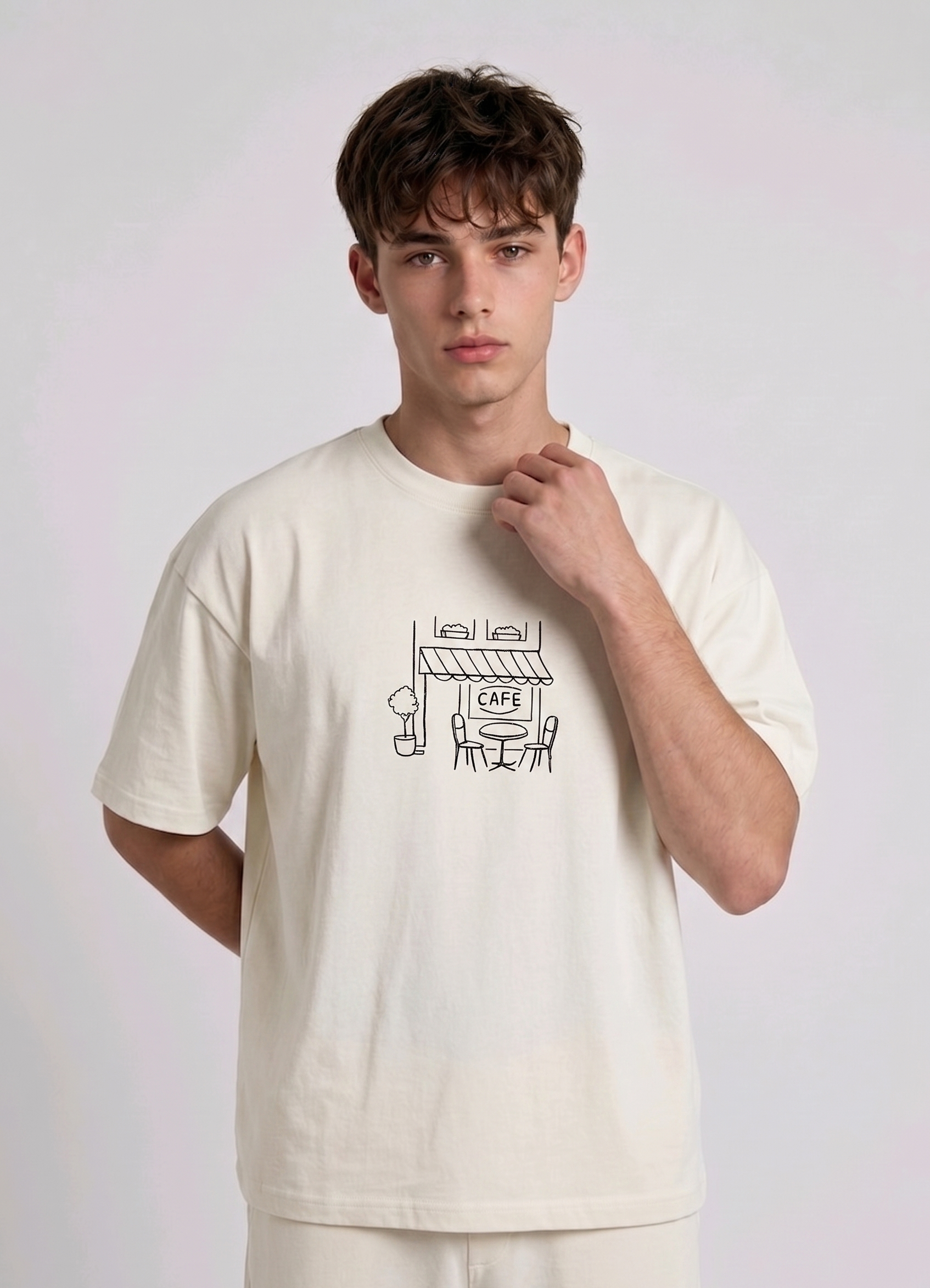Wearing a cream-colored, crew-neck t-shirt. The center of the chest features a minimalist black line-art embroidery depicting a classic cafe storefront.
