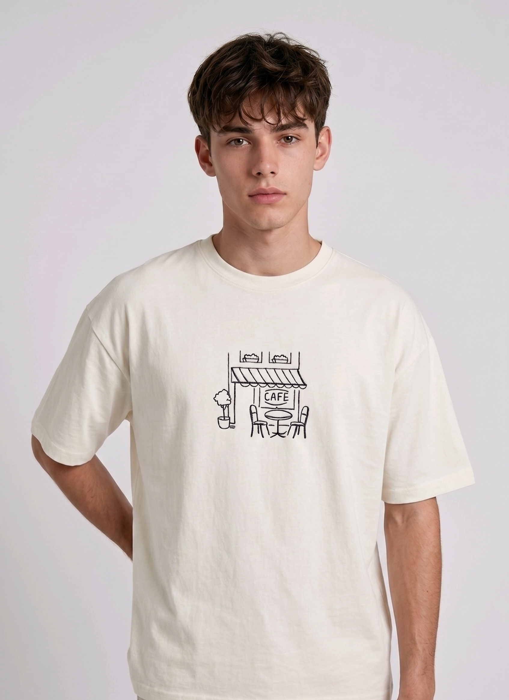 Wearing a cream-colored, crew-neck t-shirt. The center of the chest features a minimalist black line-art embroidery depicting a classic cafe storefront.
