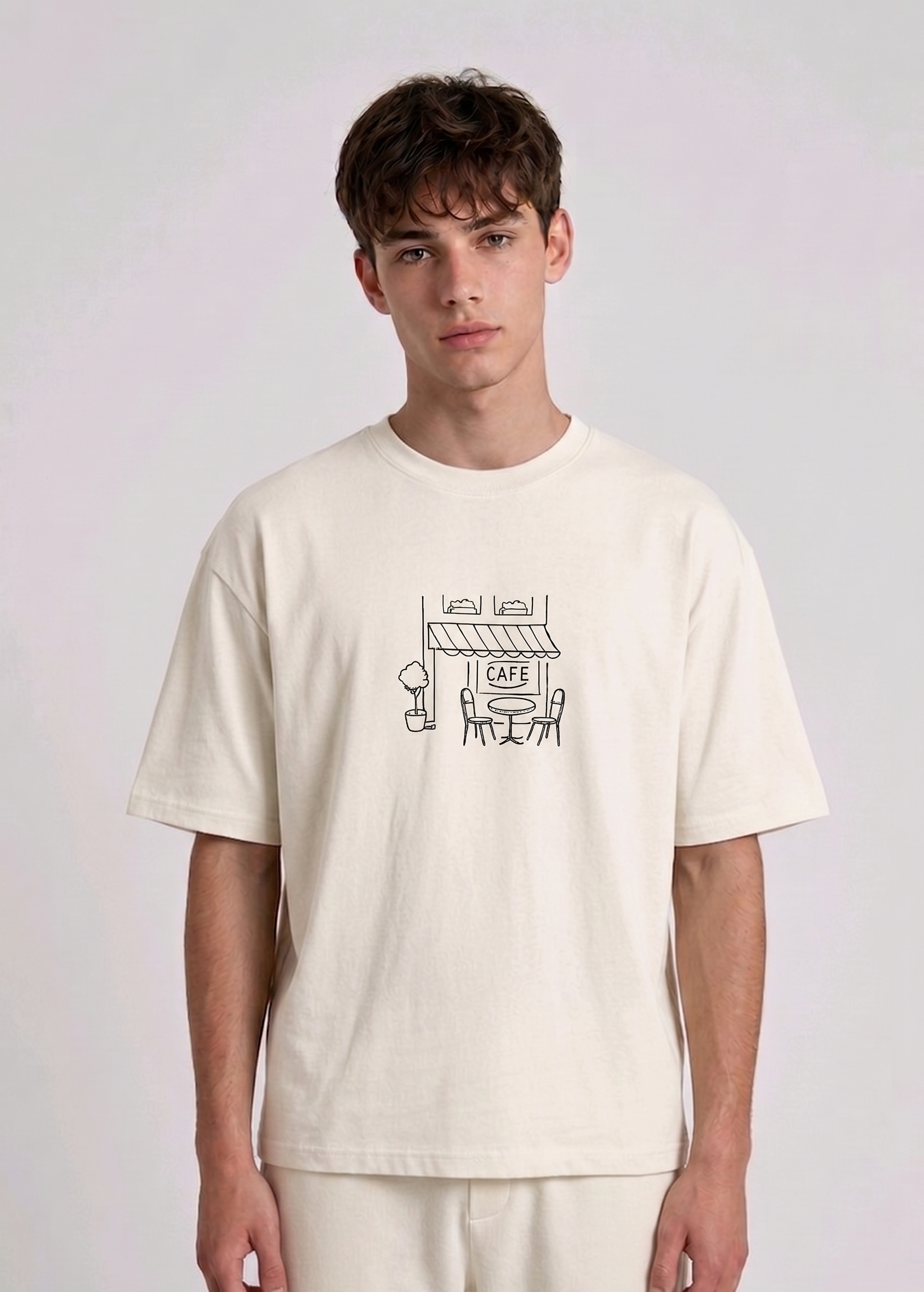 Wearing a cream-colored, crew-neck t-shirt. The center of the chest features a minimalist black line-art embroidery depicting a classic cafe storefront.
