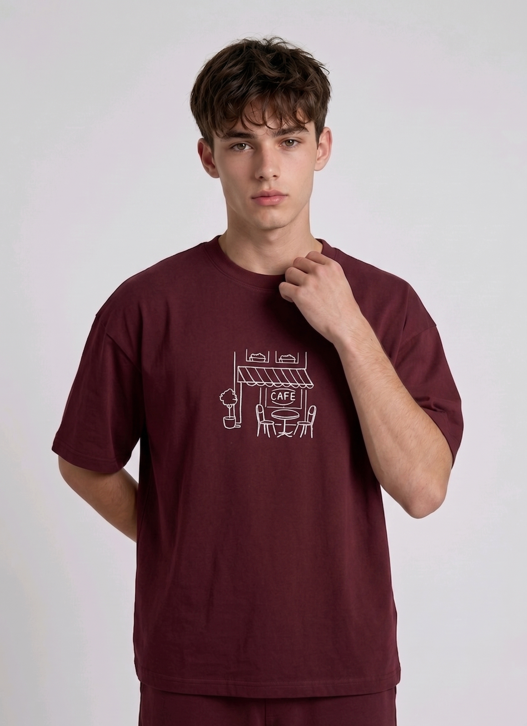 Wearing marun-colored, crew-neck t-shirt. The center of the chest features a minimalist white line-art embroidery depicting a classic cafe storefront.
