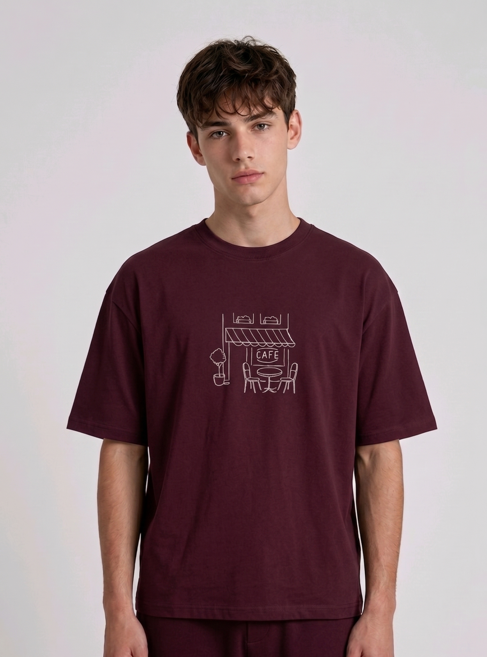 Wearing marun-colored, crew-neck t-shirt. The center of the chest features a minimalist white line-art embroidery depicting a classic cafe storefront.