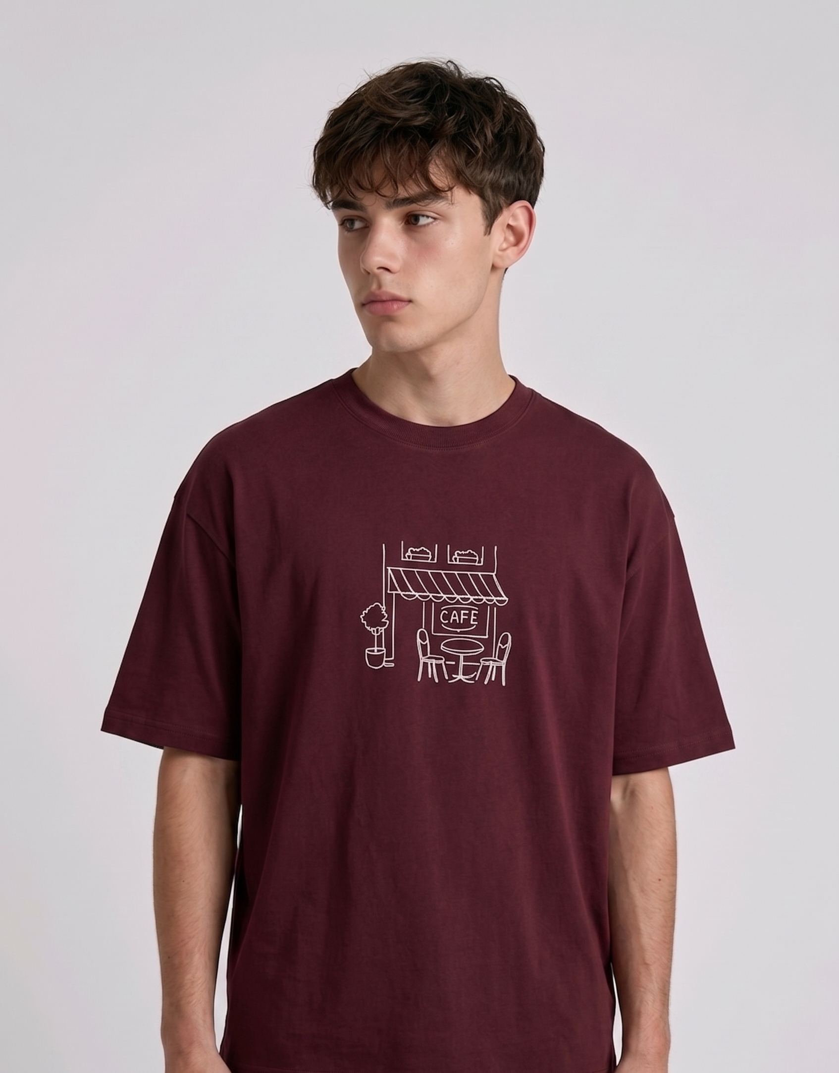 Wearing marun-colored, crew-neck t-shirt. The center of the chest features a minimalist white line-art embroidery depicting a classic cafe storefront.