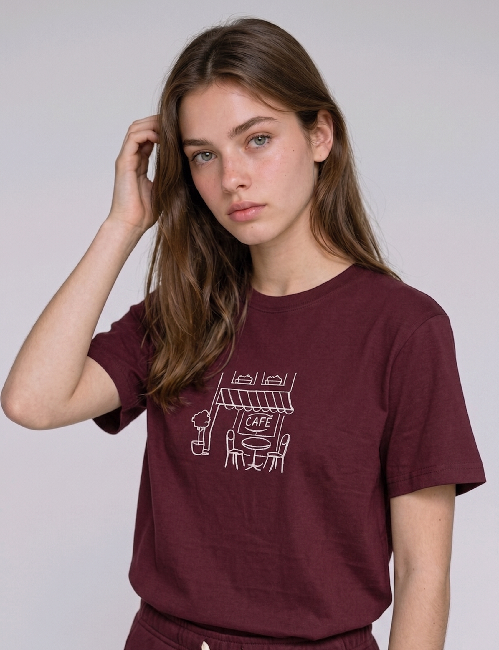 Wearing a cream-colored, crew-neck t-shirt. The center of the chest features a minimalist black line-art embroidery depicting a classic cafe storefront.

