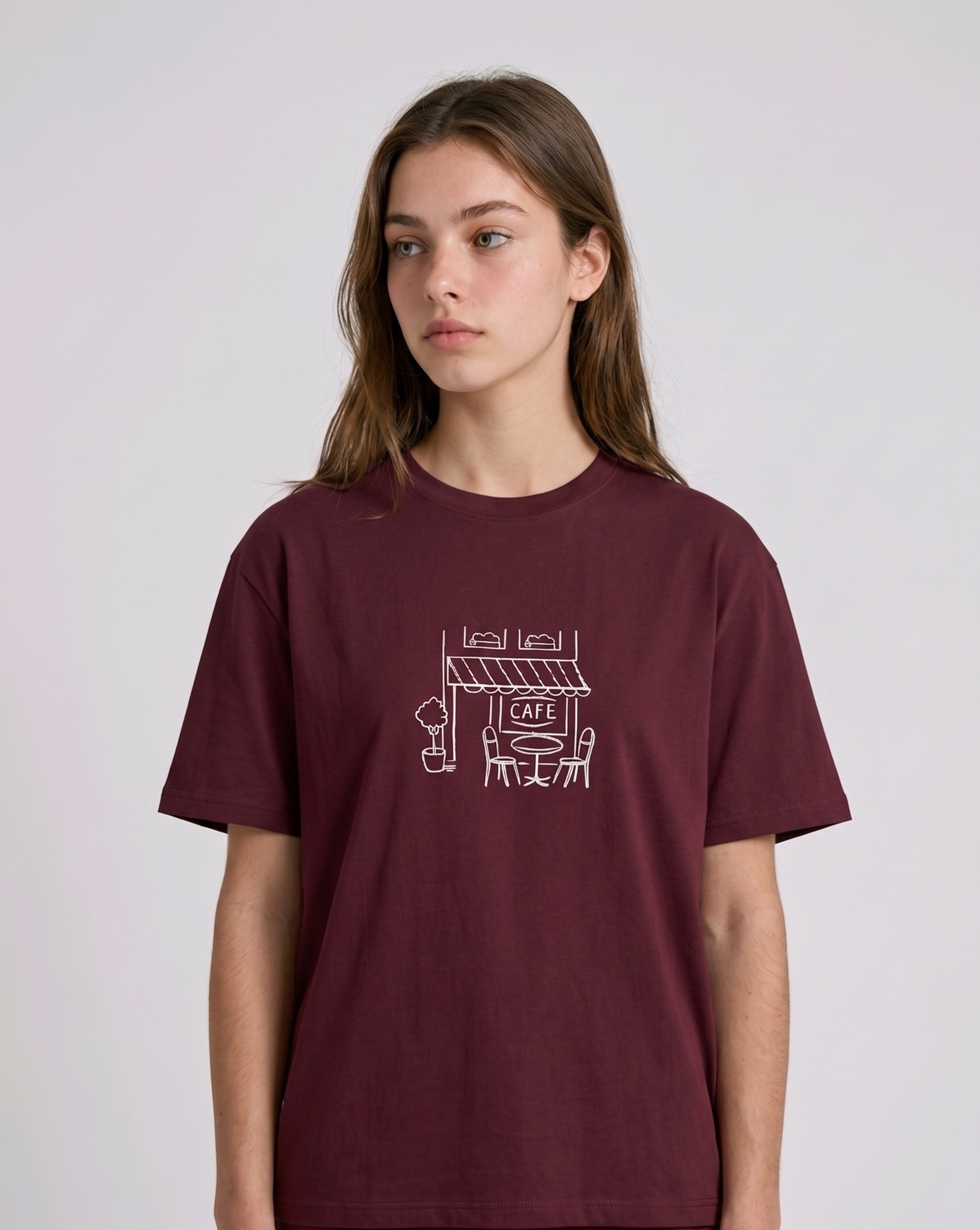 Wearing a cream-colored, crew-neck t-shirt. The center of the chest features a minimalist black line-art embroidery depicting a classic cafe storefront.

