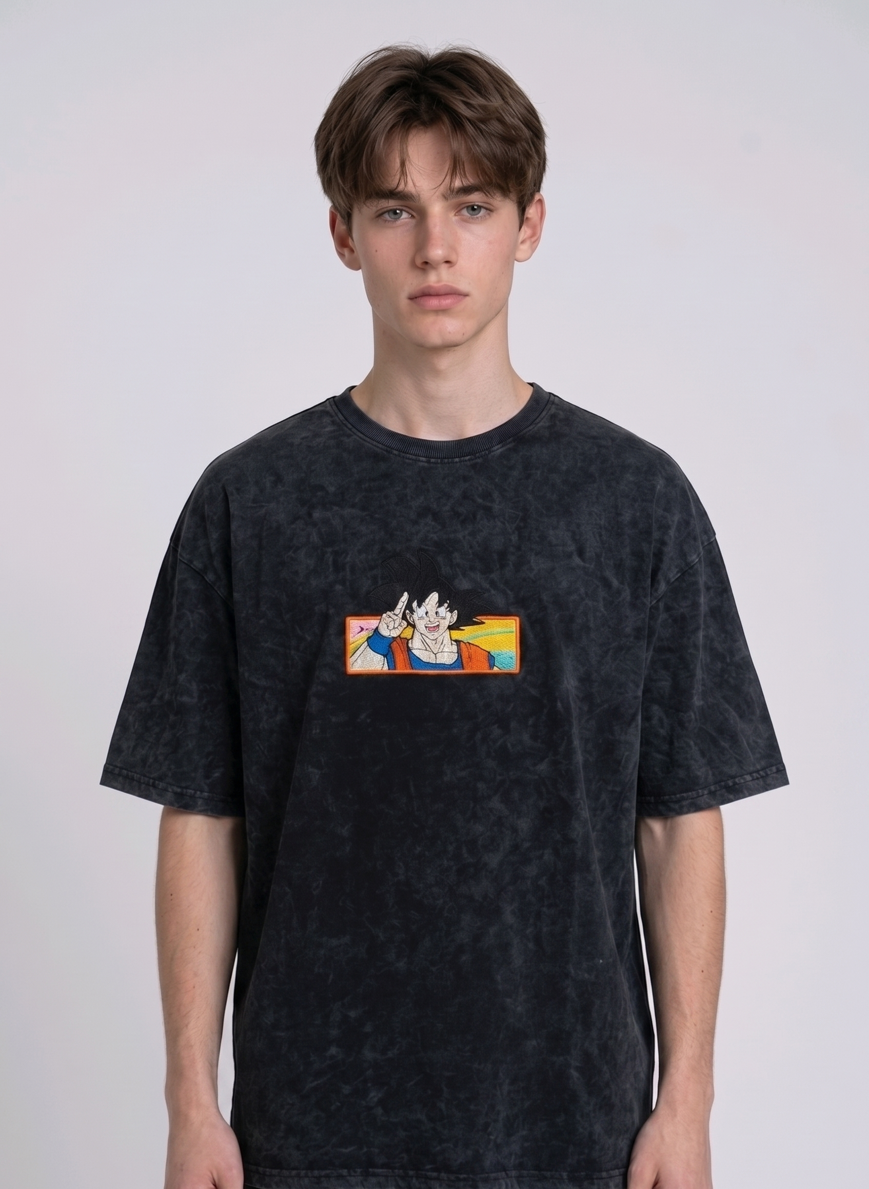 Front view of a male model with middle-parted brown hair wearing an Anetoonz dark grey mineral wash oversized t-shirt featuring a detailed, embroidered graphic of Son Goku from Dragon Ball Z giving a peace sign within a colorful framed background.