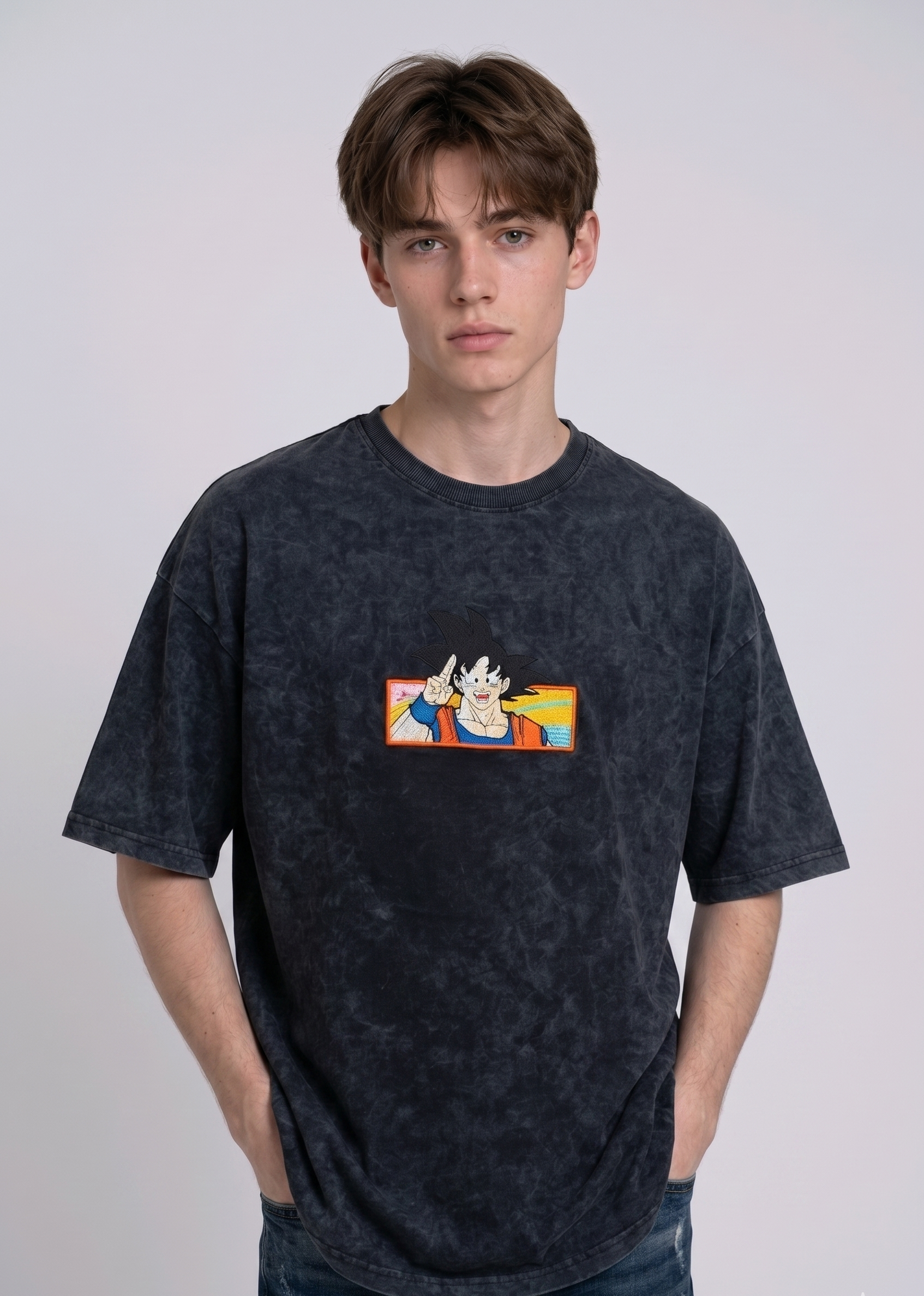 Front view of a male model with middle-parted brown hair wearing an Anetoonz dark grey mineral wash oversized t-shirt featuring a detailed, embroidered graphic of Son Goku from Dragon Ball Z giving a peace sign within a colorful framed background.