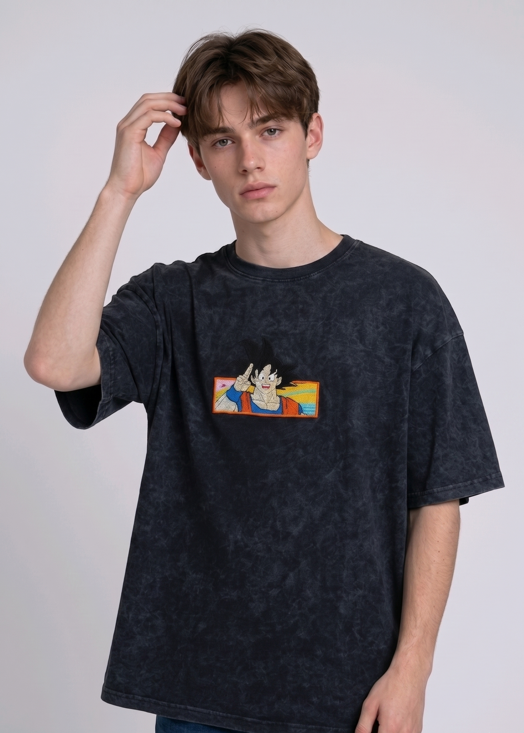 Front view of a male model with middle-parted brown hair wearing an Anetoonz dark grey mineral wash oversized t-shirt featuring a detailed, embroidered graphic of Son Goku from Dragon Ball Z giving a peace sign within a colorful framed background.