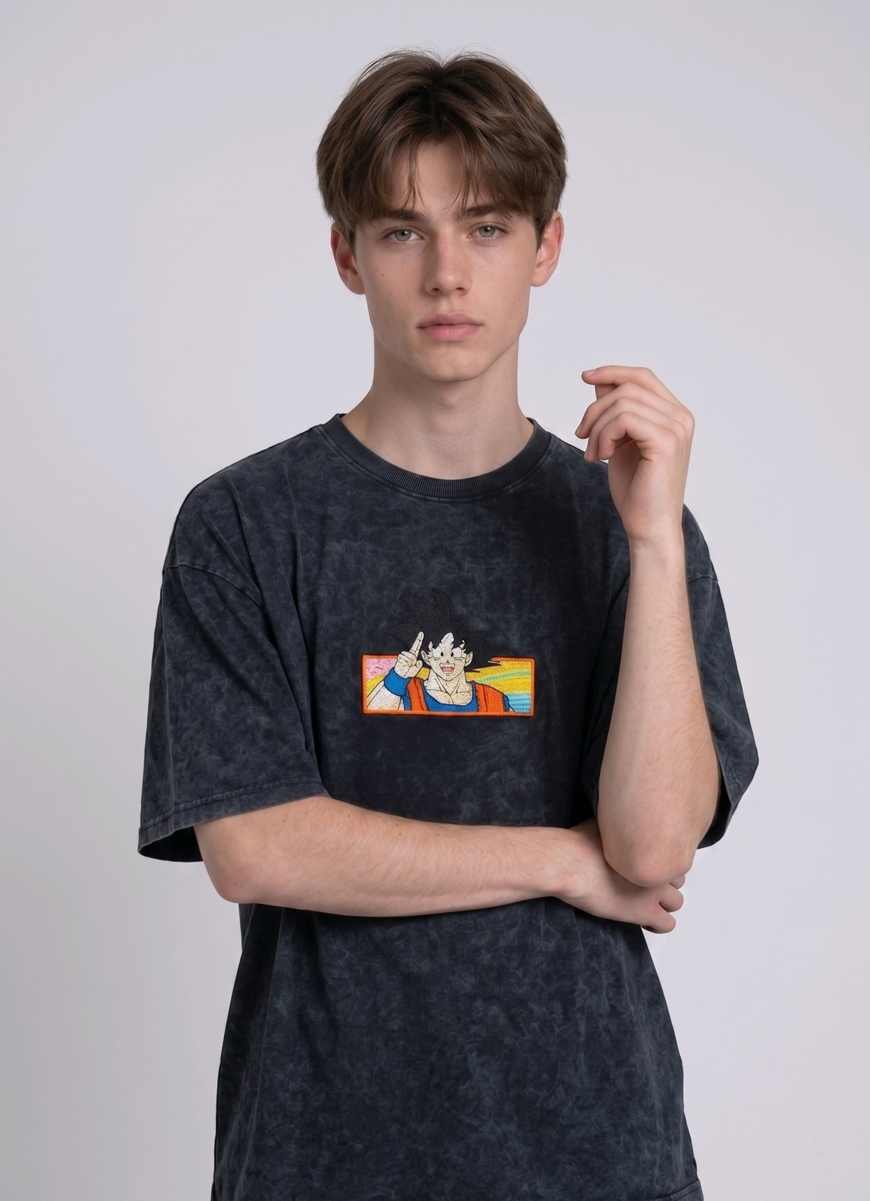 Front view of a male model with middle-parted brown hair wearing an Anetoonz dark grey mineral wash oversized t-shirt featuring a detailed, embroidered graphic of Son Goku from Dragon Ball Z giving a peace sign within a colorful framed background.