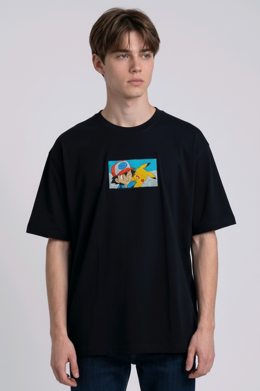 He is wearing a dark, heavyweight cotton t-shirt with a detailed, colorful embroidered patch on the chest featuring anime characters Ash Ketchum (winking) and Pikachu.