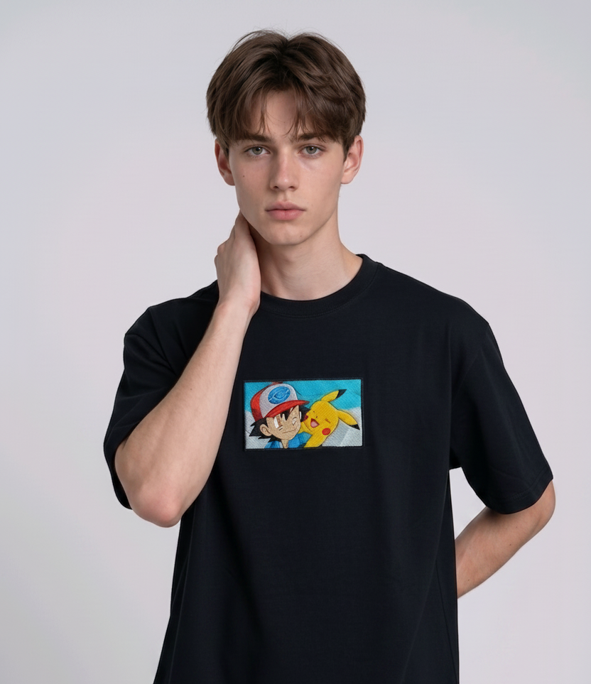 He is wearing a dark, heavyweight cotton t-shirt with a detailed, colorful embroidered patch on the chest featuring anime characters Ash Ketchum (winking) and Pikachu.