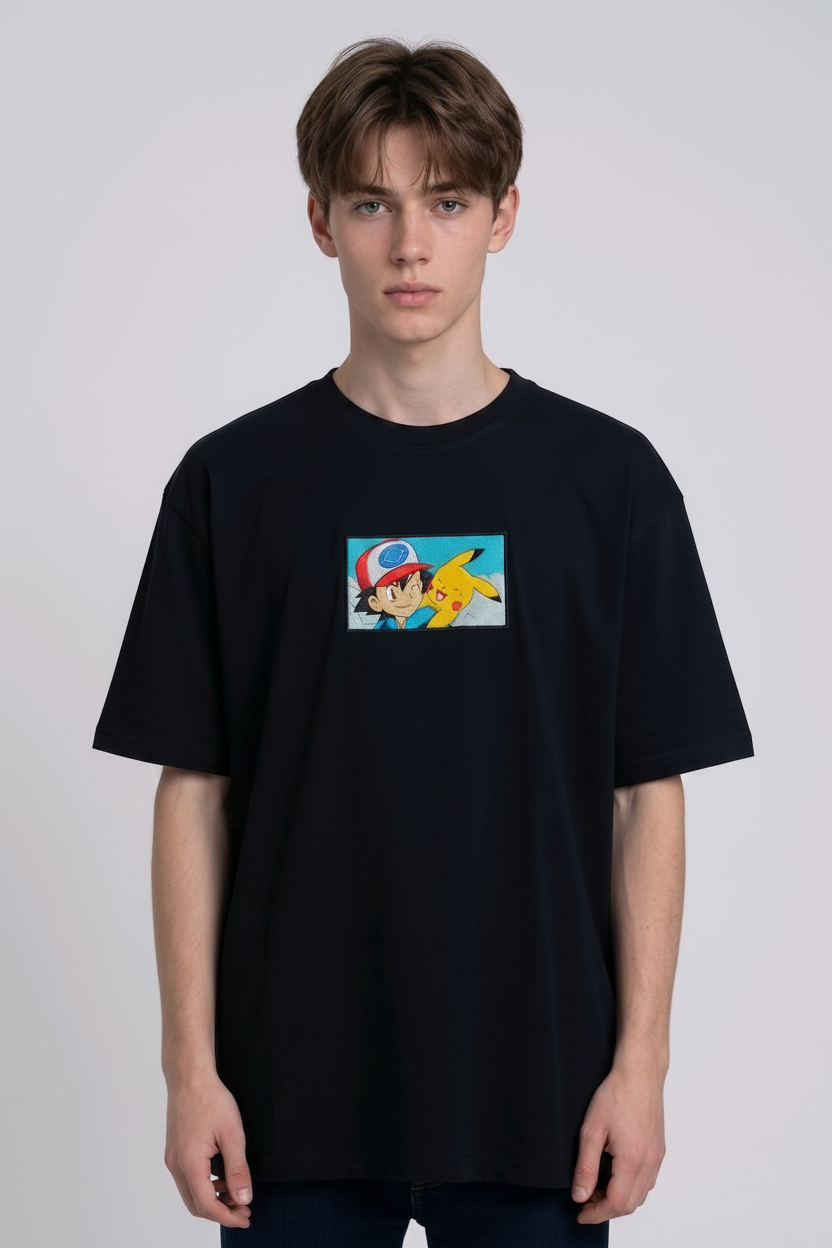 He is wearing a dark, heavyweight cotton t-shirt with a detailed, colorful embroidered patch on the chest featuring anime characters Ash Ketchum (winking) and Pikachu.