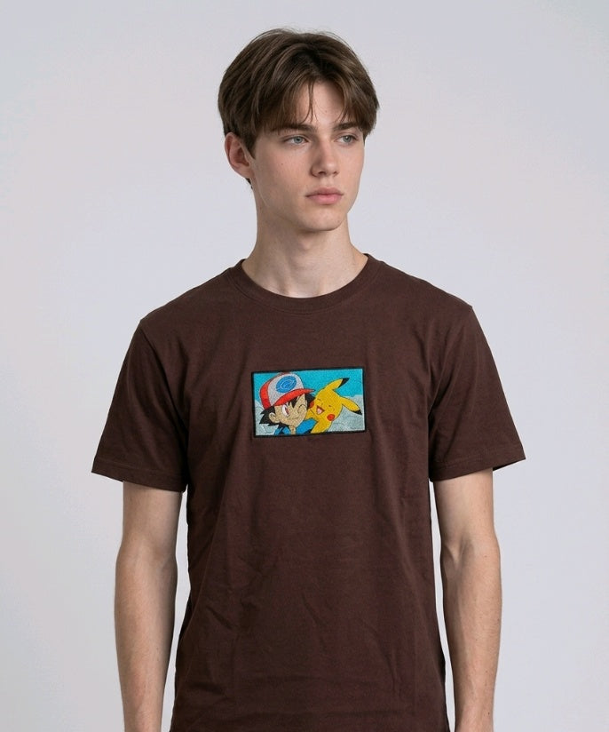 He is wearing , heavyweight cotton t-shirt with a detailed, colorful embroidered patch on the chest featuring anime characters Ash Ketchum (winking) and Pikachu.