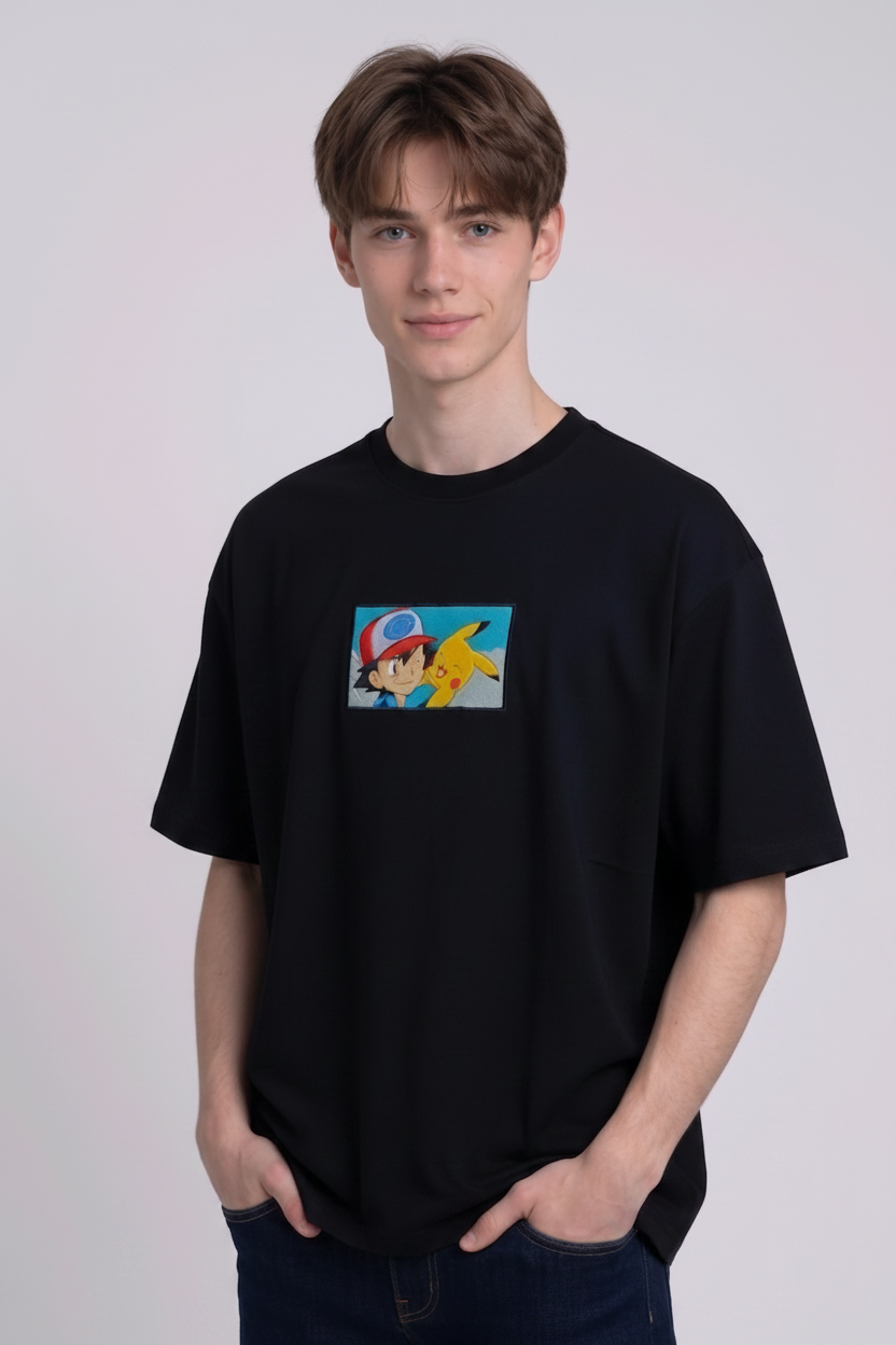 He is wearing a dark, heavyweight cotton t-shirt with a detailed, colorful embroidered patch on the chest featuring anime characters Ash Ketchum (winking) and Pikachu.